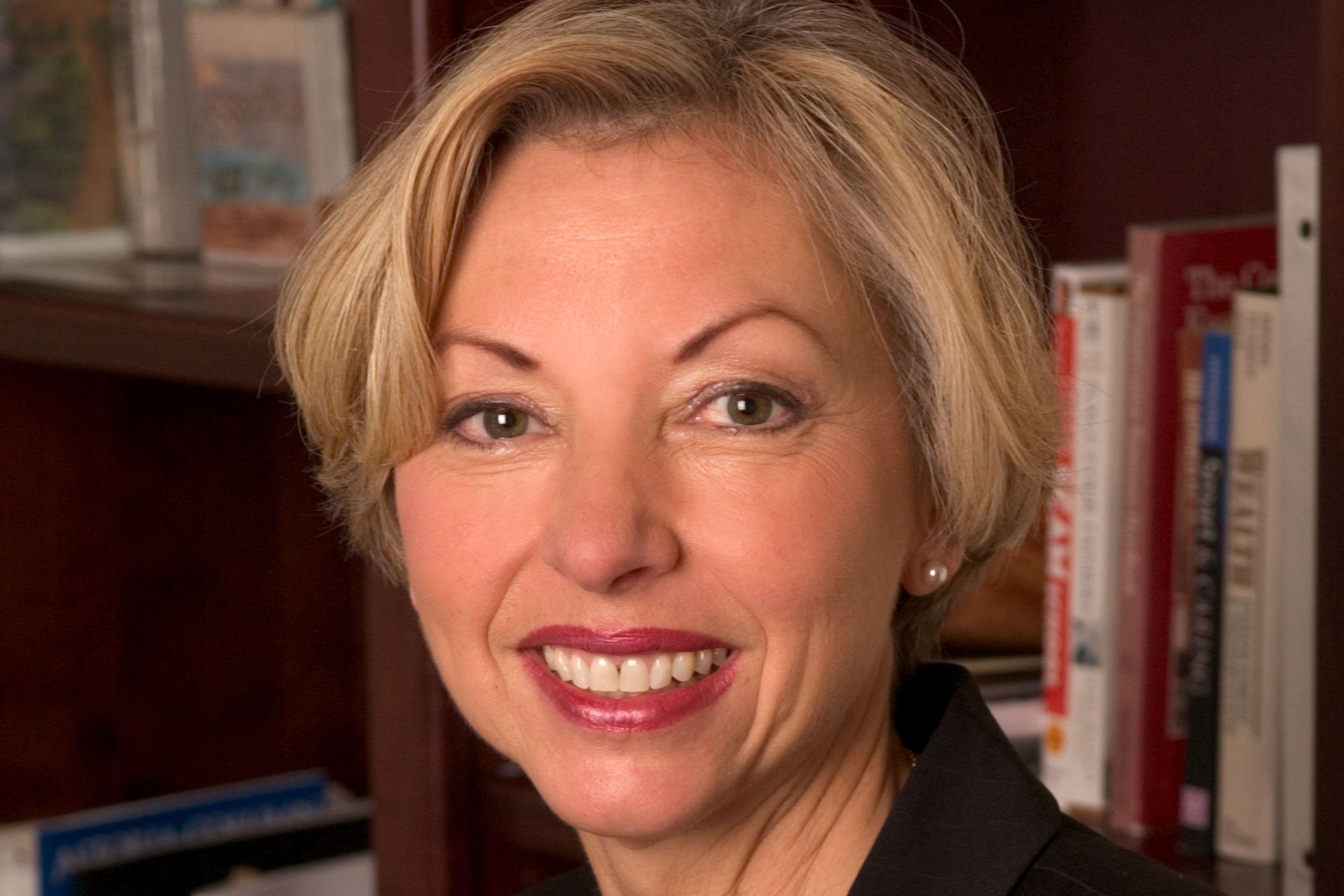 A headshot of Nancy Hales smiling in front of a bookcase while wearing a black blazer, white shirt and necklace.