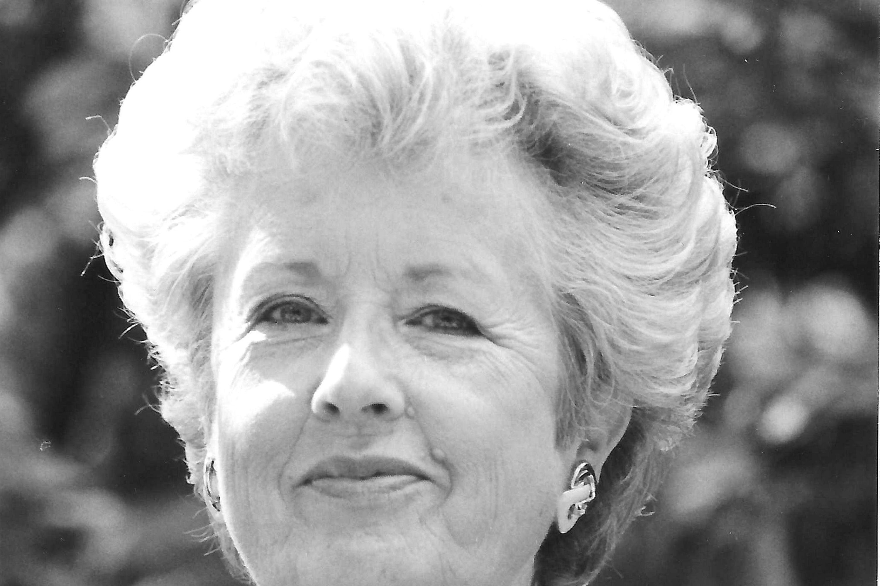 A black and white portrait of Mary Granger smirking in a floral print dress with earings and a necklace on.