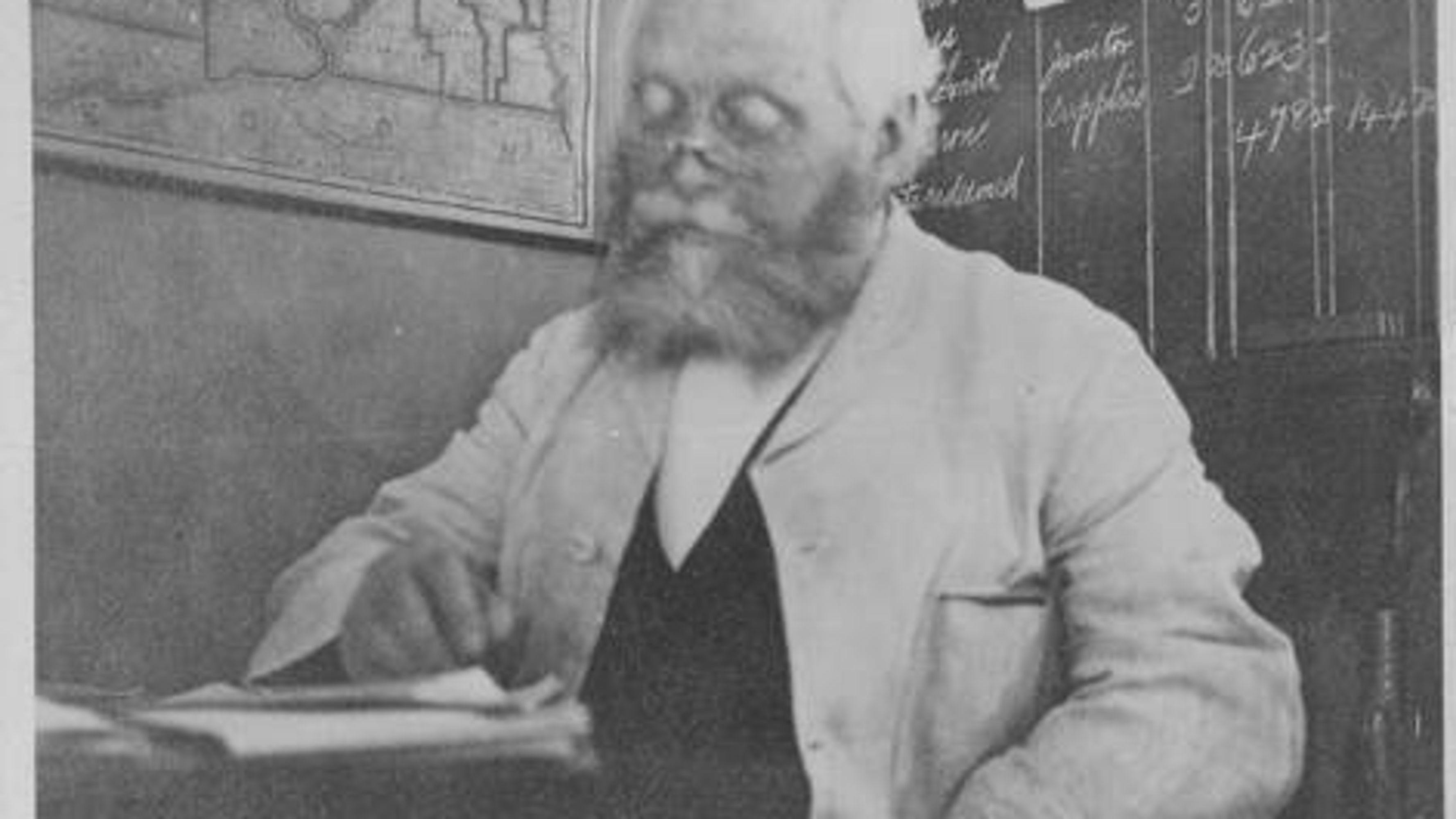 An old photograph of an old professor reading notes by a chalkboard