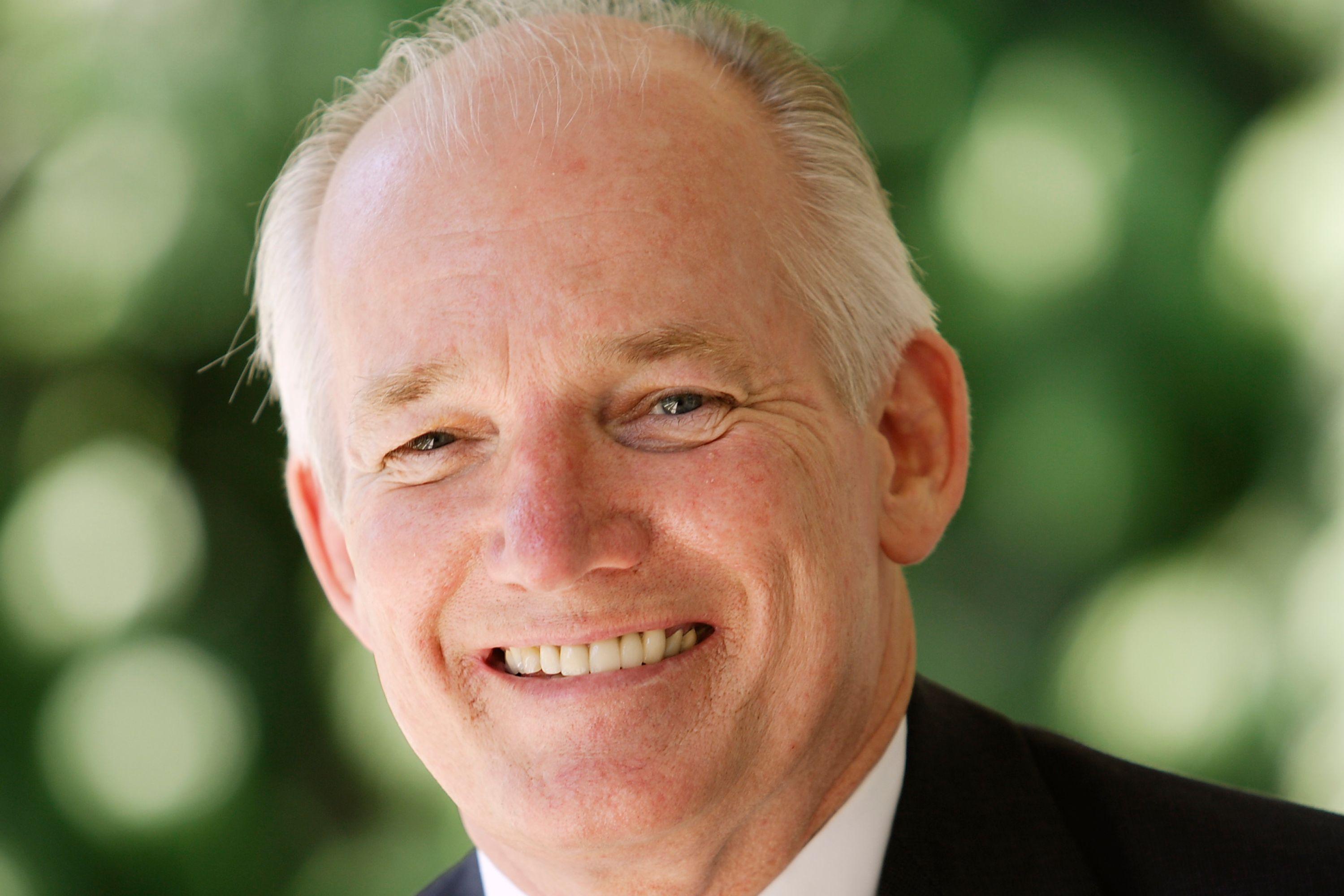 A headshot of Richard Melching smiling while wearing a black suit jacket with a blue and white striped tie.