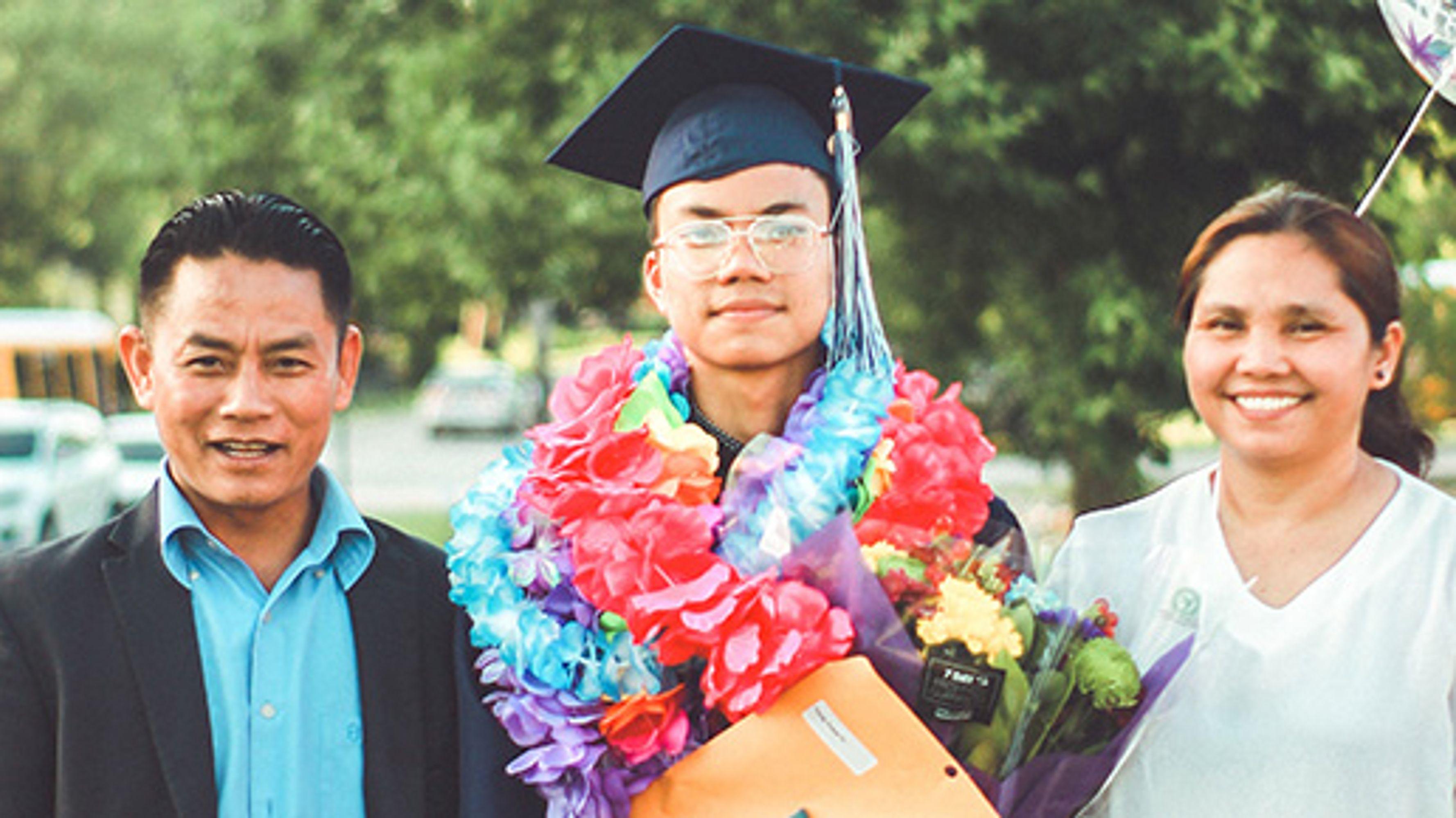 A graduate in a cap and many leis standing next to a man and a woman