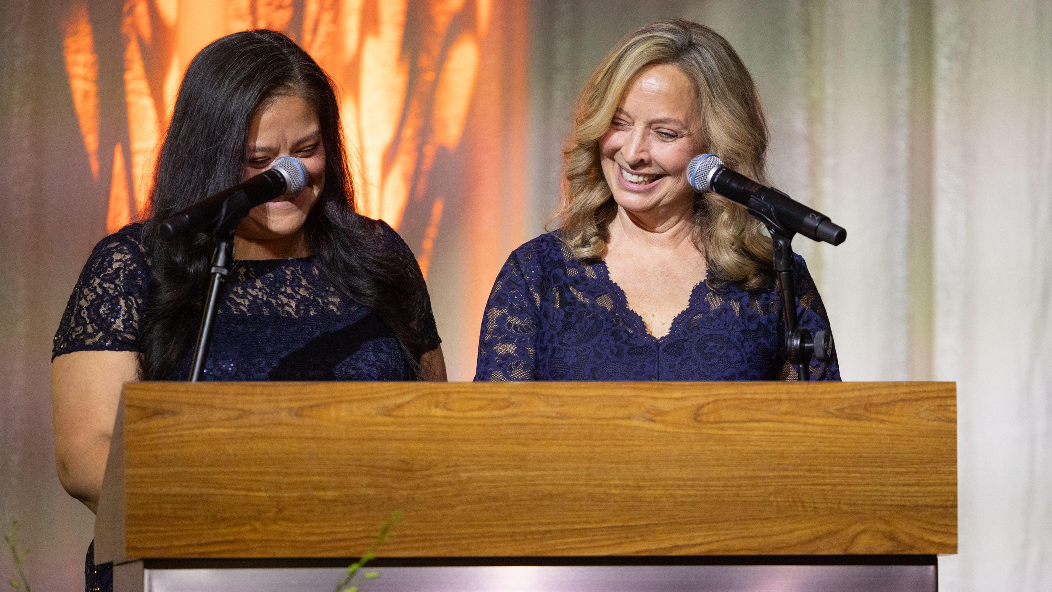 Two women in navy blue lace dresses standing behind a podium smiling and talking