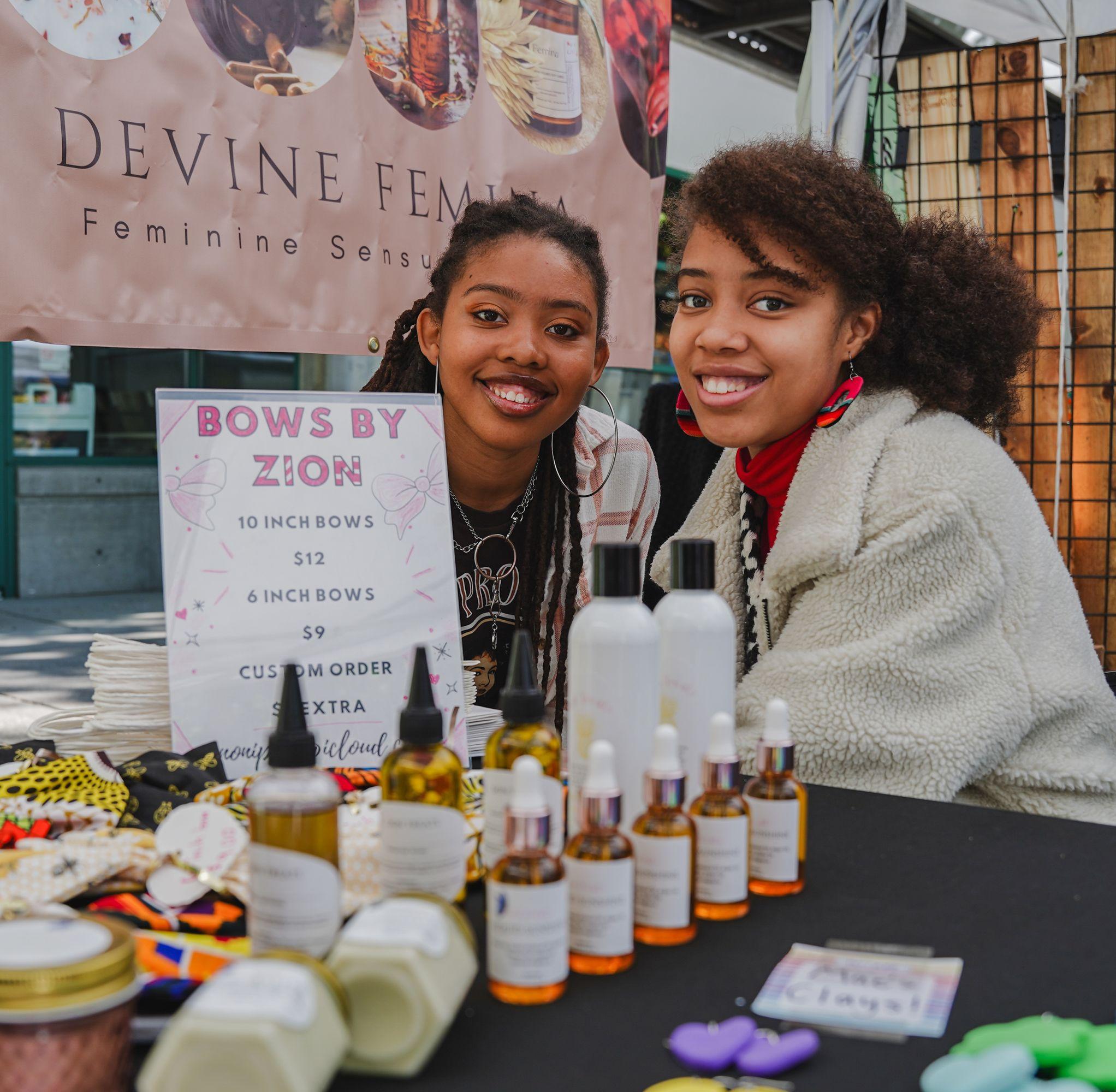 A pair of young African American women sit smiling behind a table displaying their beauty products at the Vancouver Farmers Market.