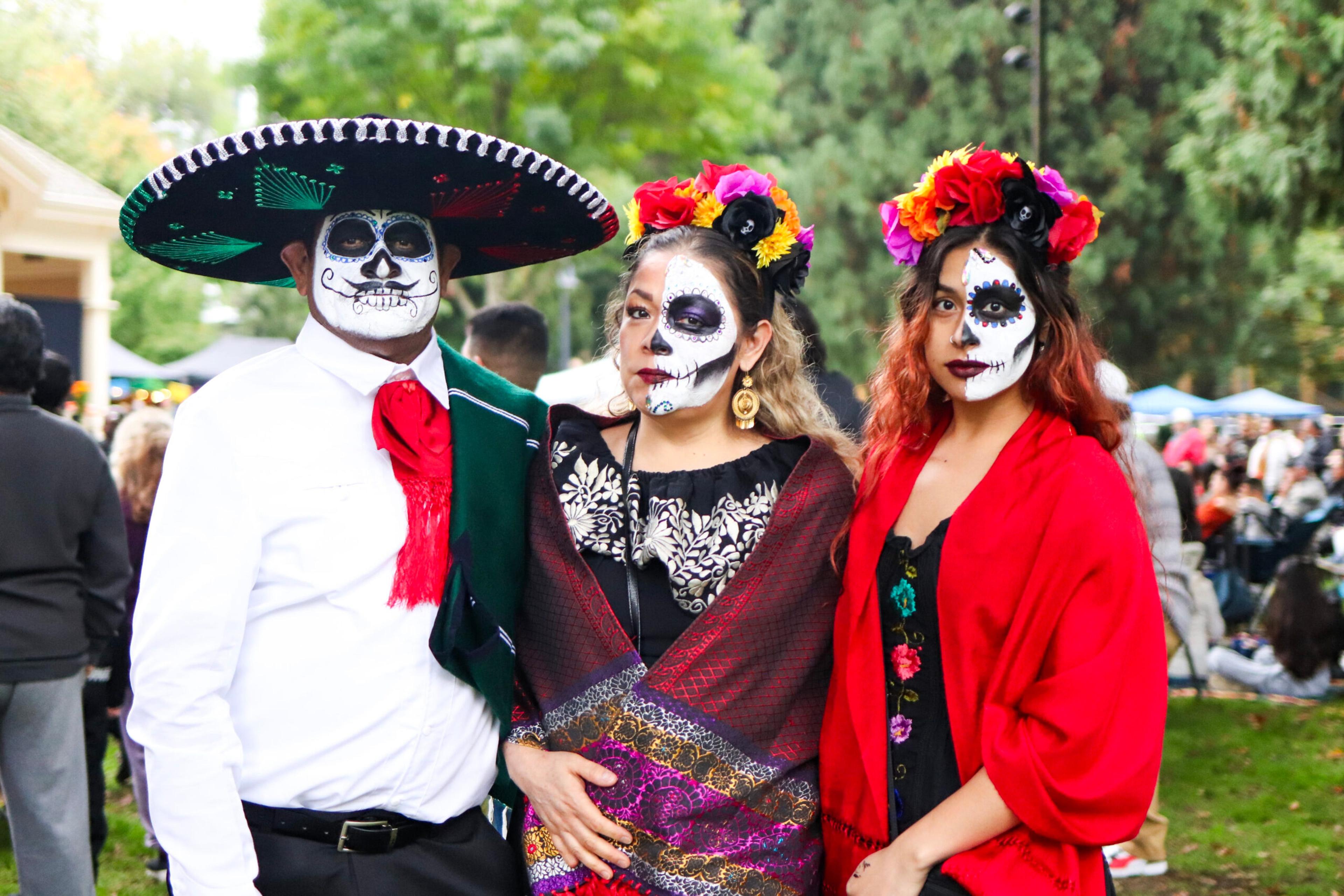 Three people dressed in traditional Dia de Muertos suits and dresses with painted faces.