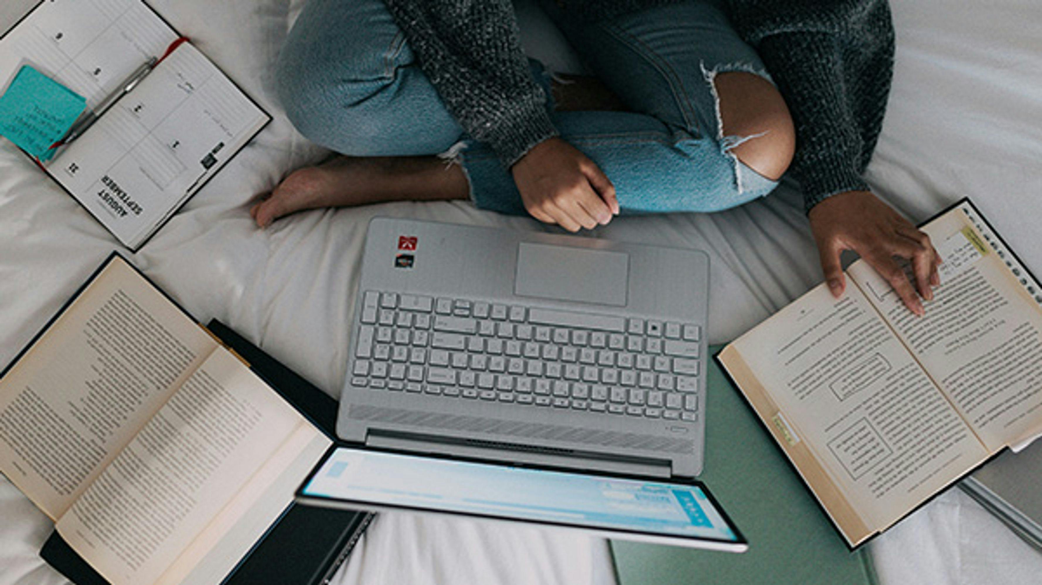 Young person sitting on a bed surrounded by books, a planner and a laptop