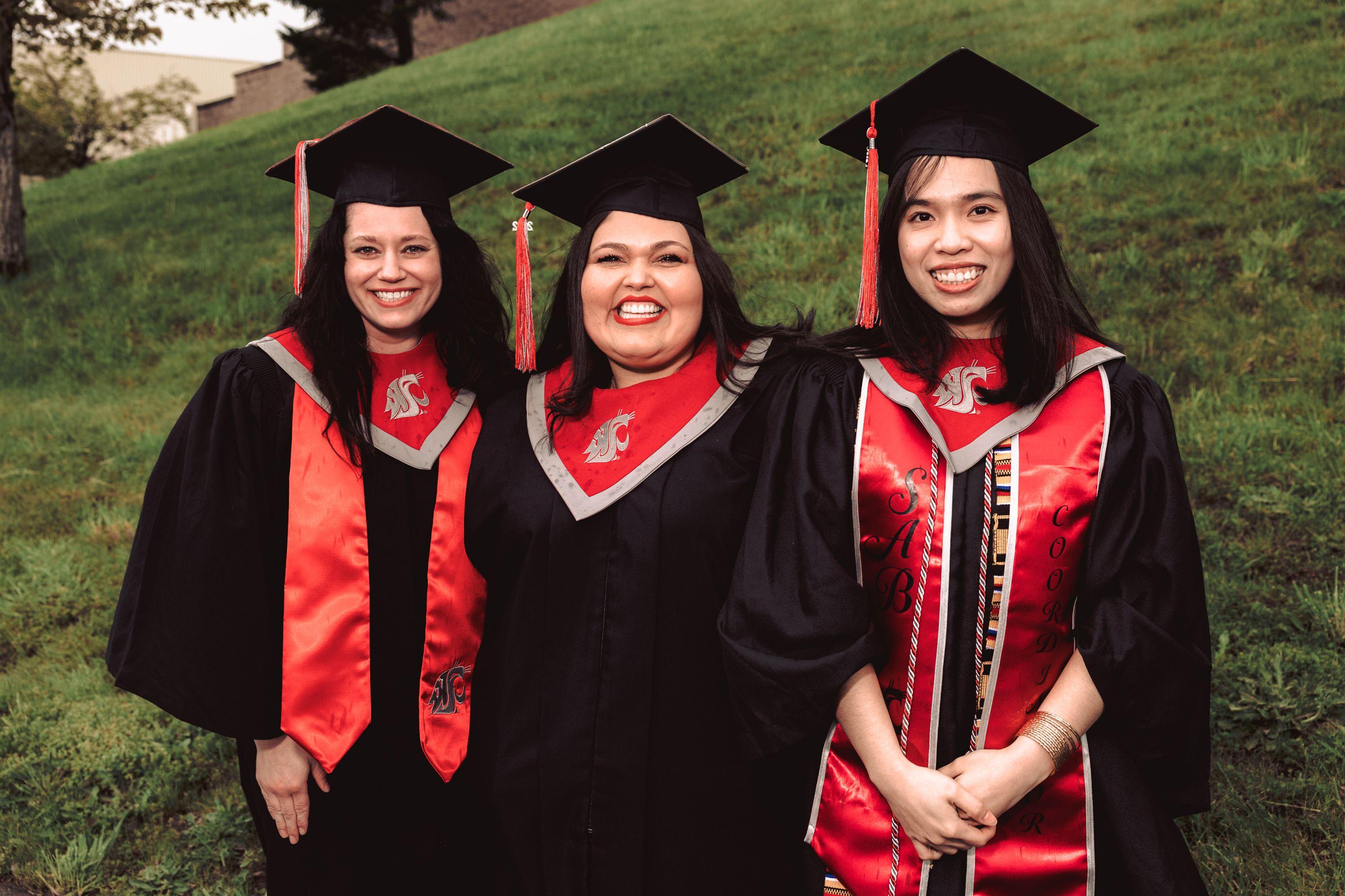 Three women of varying ethnicity smiling in their caps and gowns after a WSU Vancouver commencement ceremony.