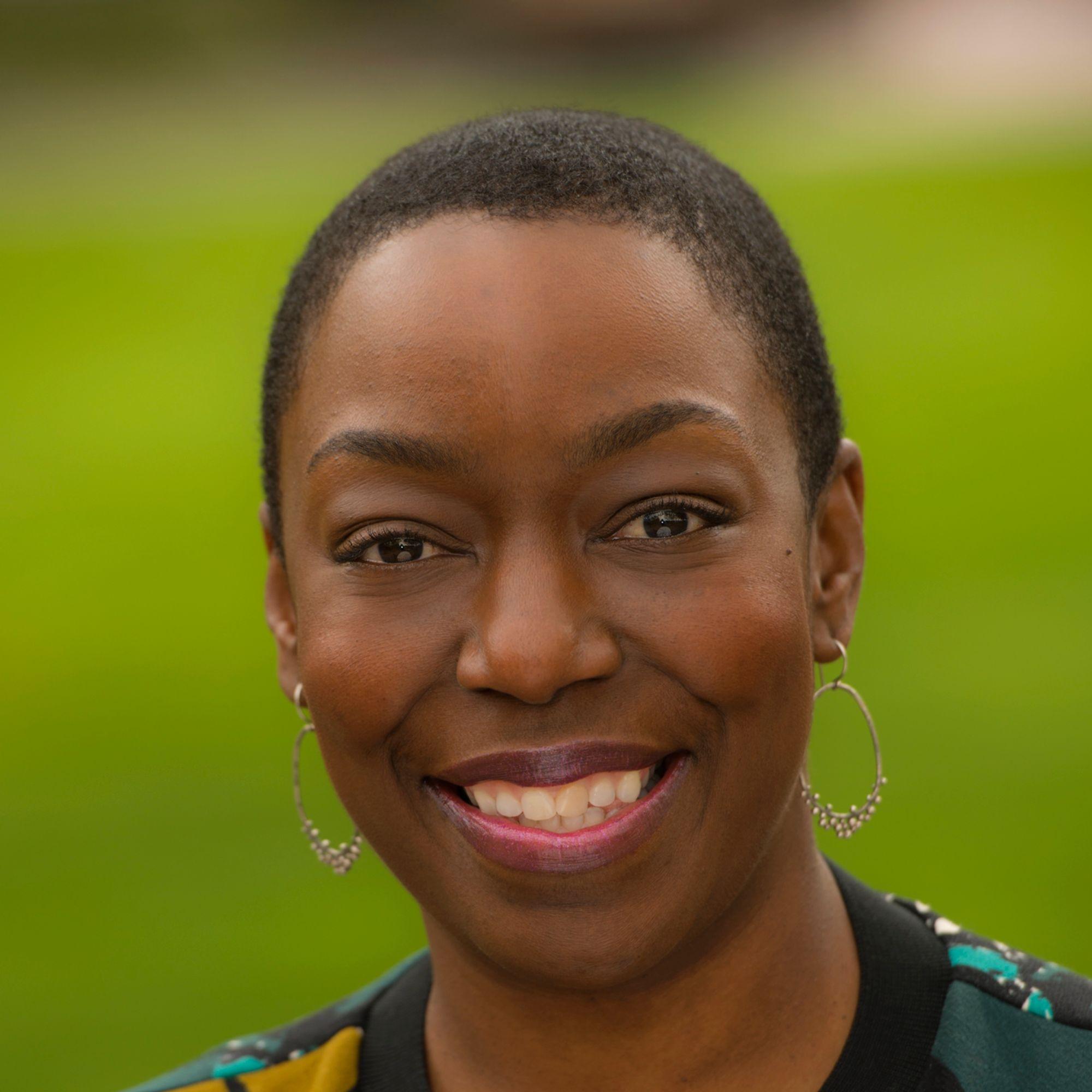 A Black woman with a shaved head and a patterned top smiling at the camera