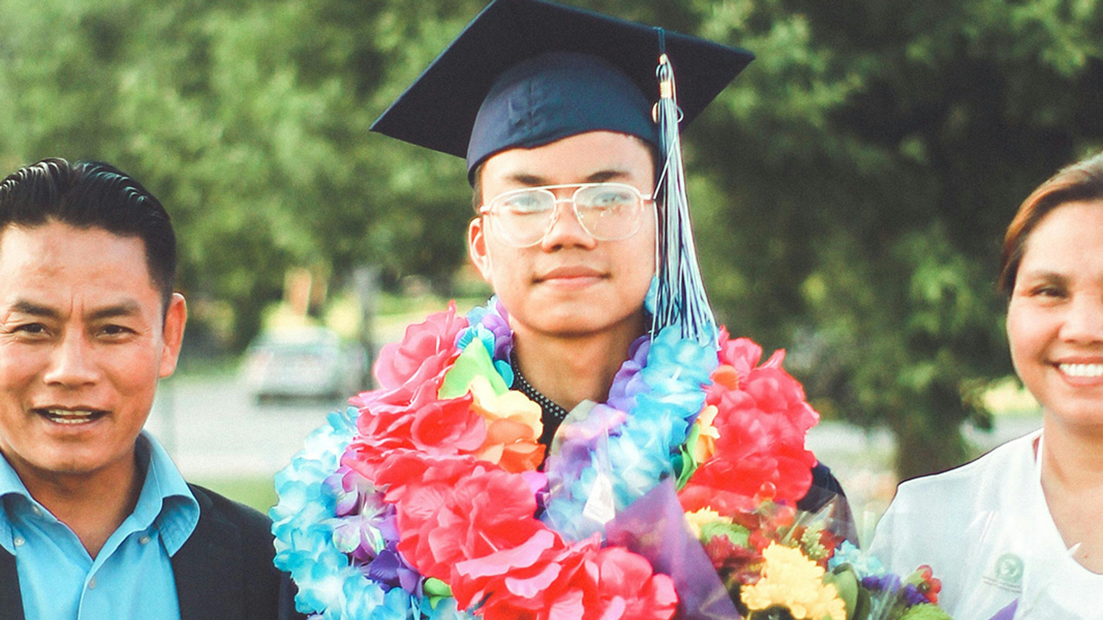 A graduate in a cap and many leis standing next to a man and a woman