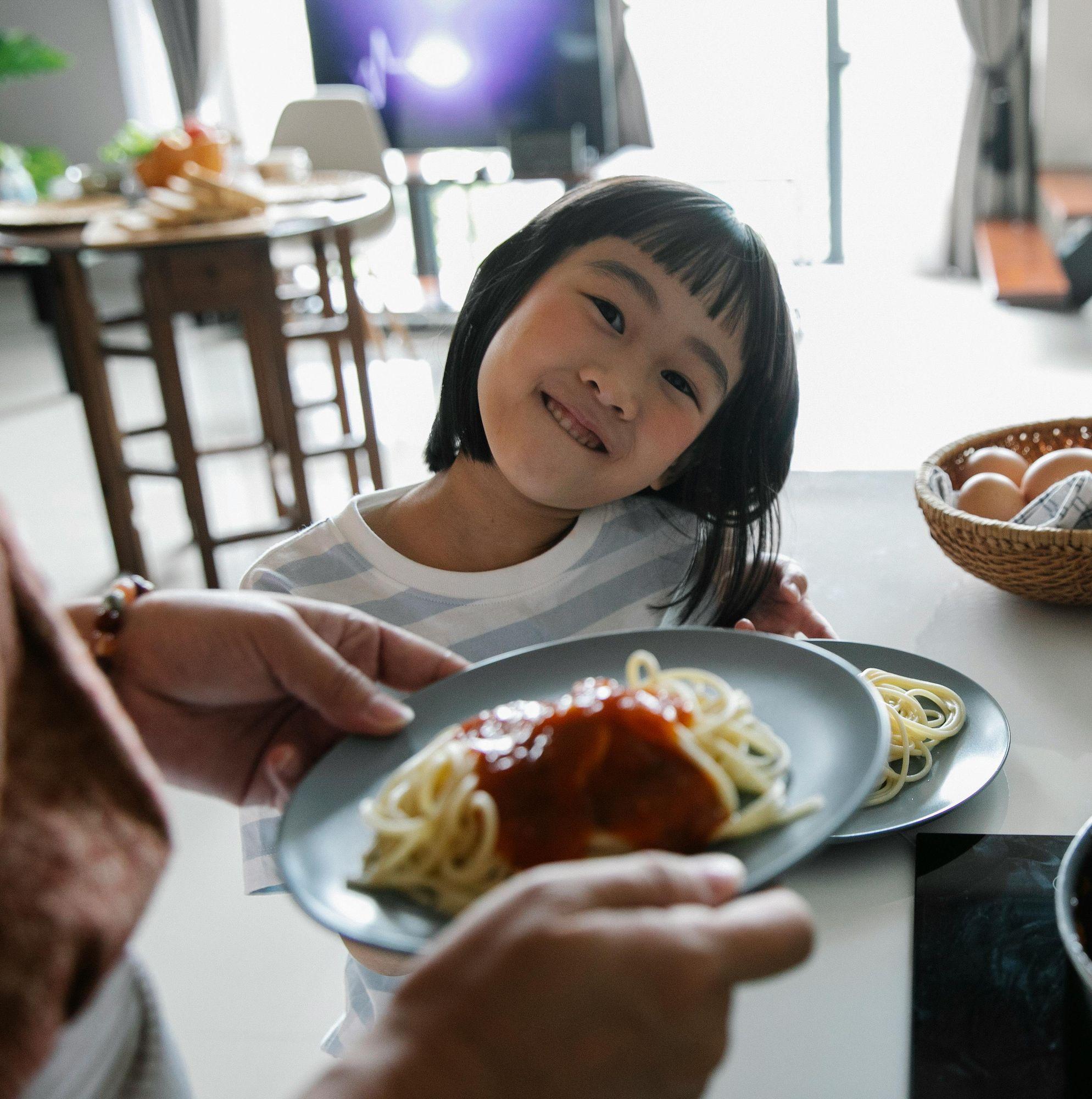 An Asian girl smiles at the camera as somone out of frame serves her a plate of spaghetti.